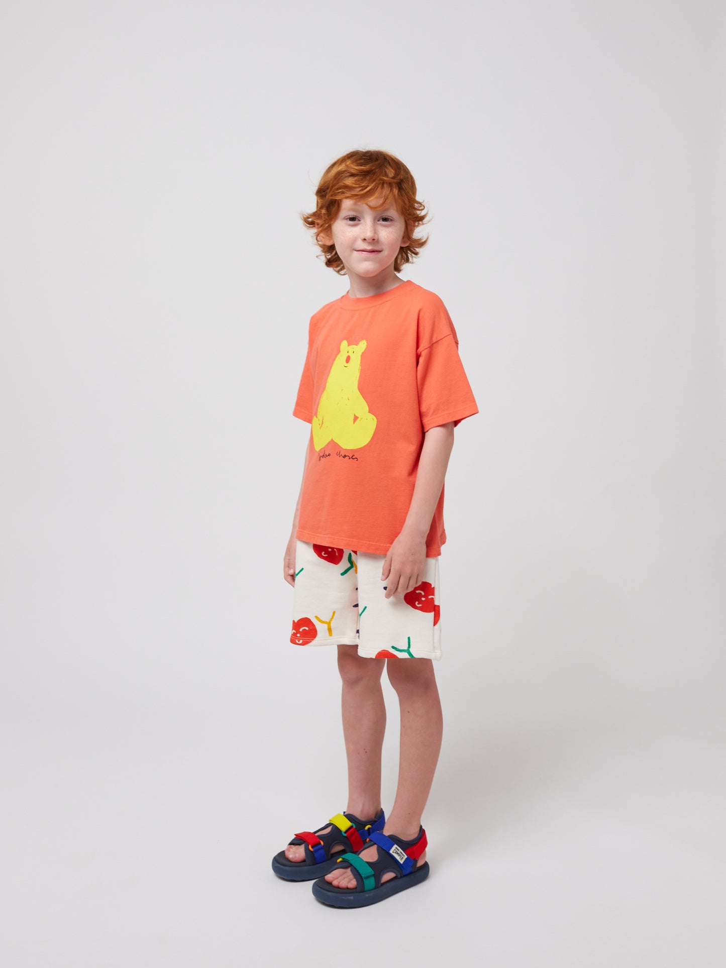 A child with curly red hair wears the coral Bobo Choses T-shirt with white shorts featuring a cherry print. They are also wearing multicoloured sporty sandals.