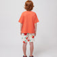 A child stands with their back facing the camera, wearing the coral T-shirt and white shorts with a playful cherry print. Their curly red hair is visible, along with multicoloured sandals.