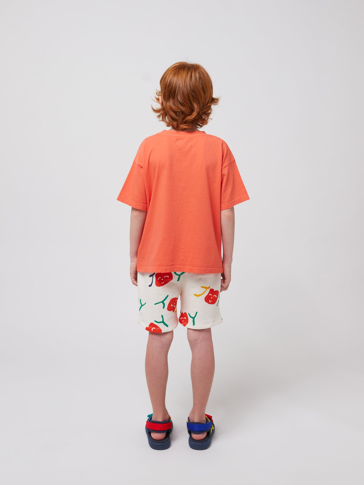 A child stands with their back facing the camera, wearing the coral T-shirt and white shorts with a playful cherry print. Their curly red hair is visible, along with multicoloured sandals.