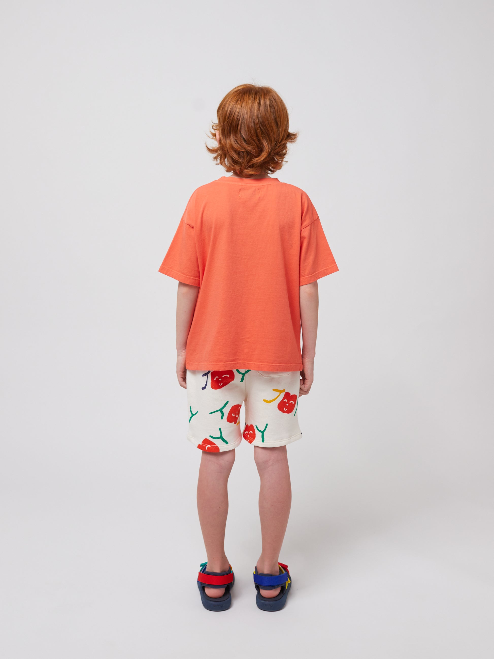 A child stands with their back facing the camera, wearing the coral T-shirt and white shorts with a playful cherry print. Their curly red hair is visible, along with multicoloured sandals.