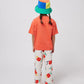 The child is standing with their back facing the camera, showing the solid coral colour of the T-shirt’s back. The bucket hat’s multicolour patchwork design is also visible from this angle.