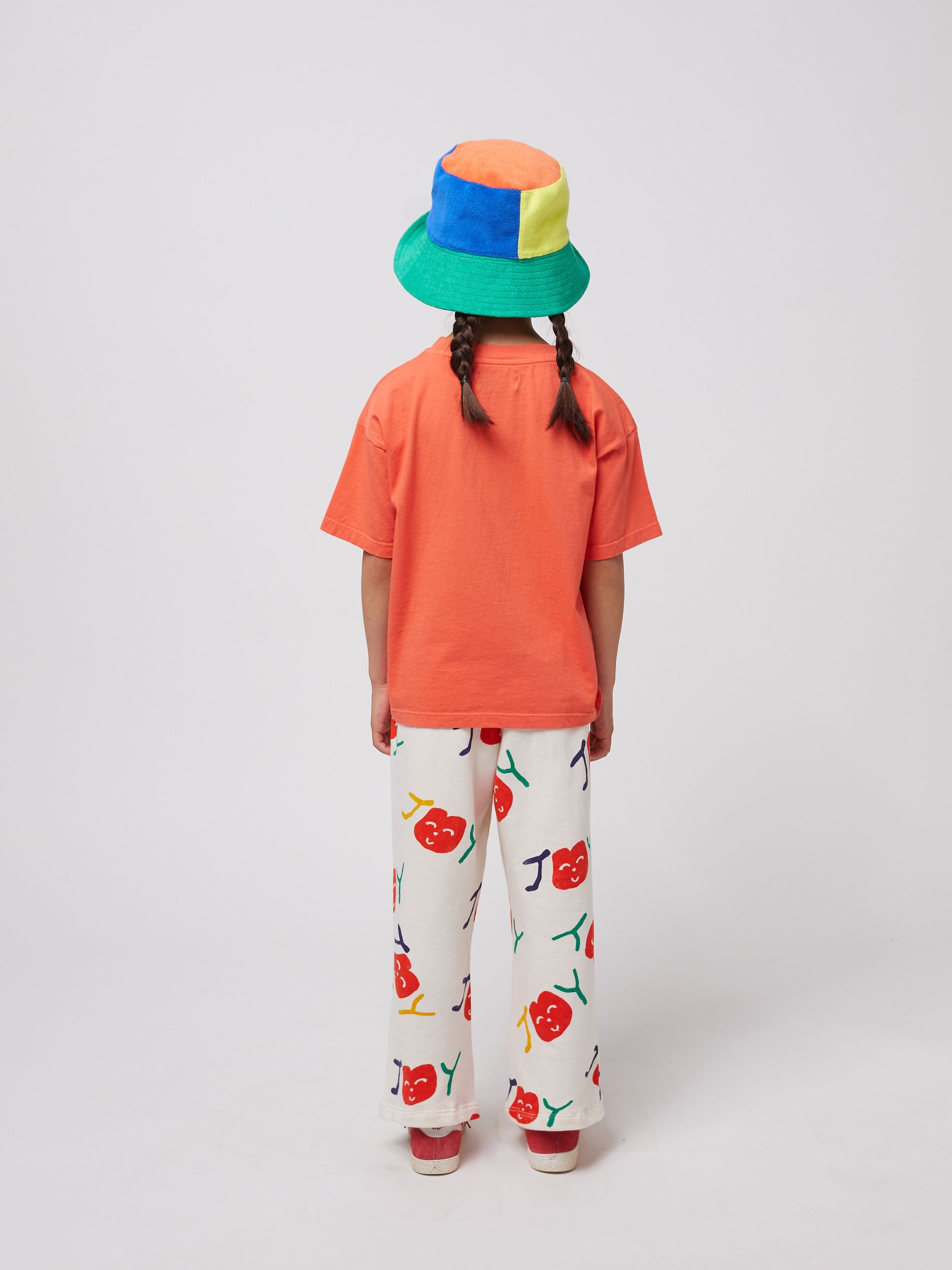 The child is standing with their back facing the camera, showing the solid coral colour of the T-shirt’s back. The bucket hat’s multicolour patchwork design is also visible from this angle.