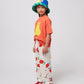 A side view of the child wearing the coral T-shirt, cherry-printed trousers, and a patchwork bucket hat. The oversized fit of the T-shirt drapes loosely.