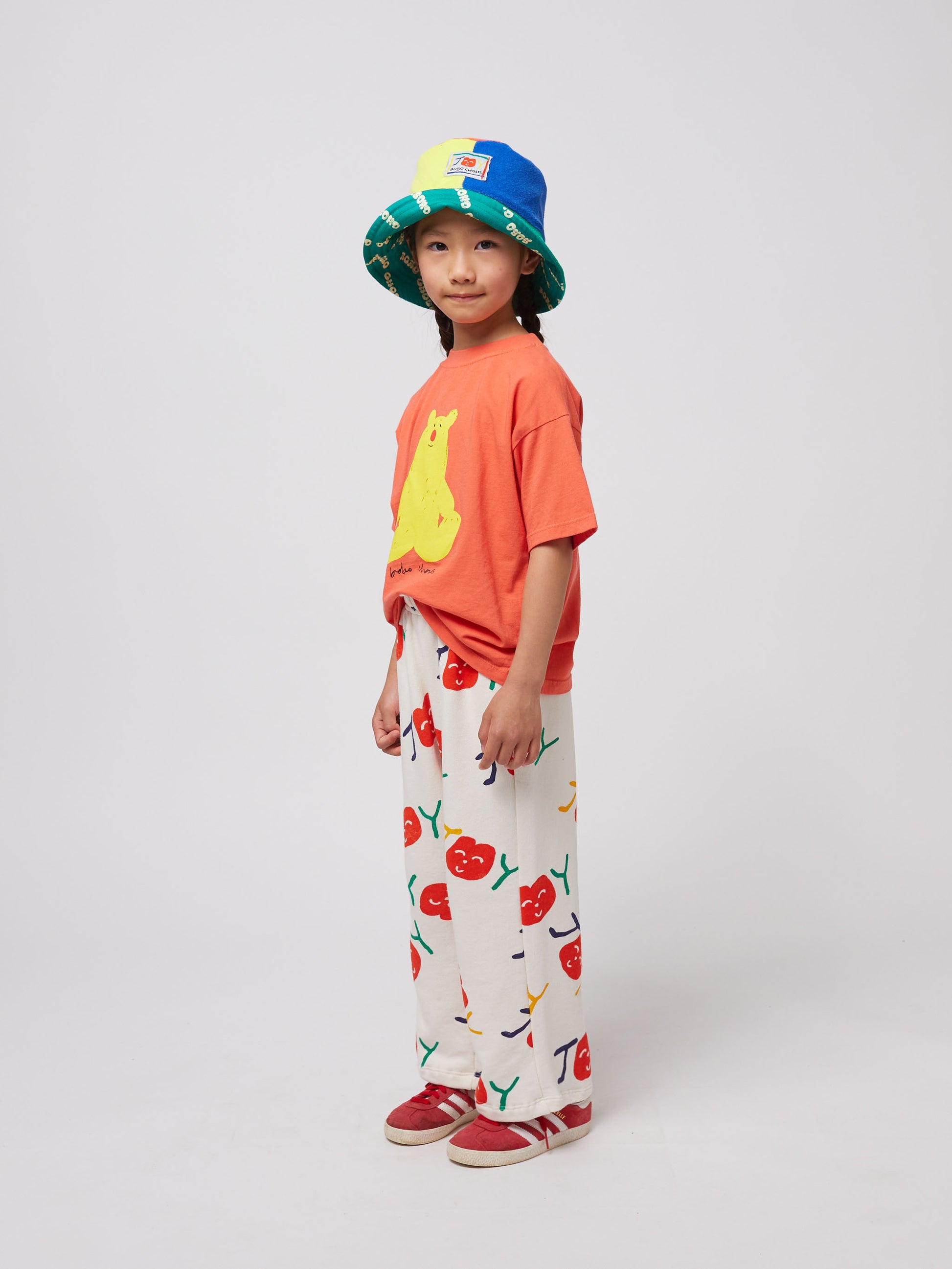 A side view of the child wearing the coral T-shirt, cherry-printed trousers, and a patchwork bucket hat. The oversized fit of the T-shirt drapes loosely.