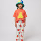 The child is standing against a plain background, dressed in the coral T-shirt, wide-leg cream trousers with a playful cherry print, and red sneakers.