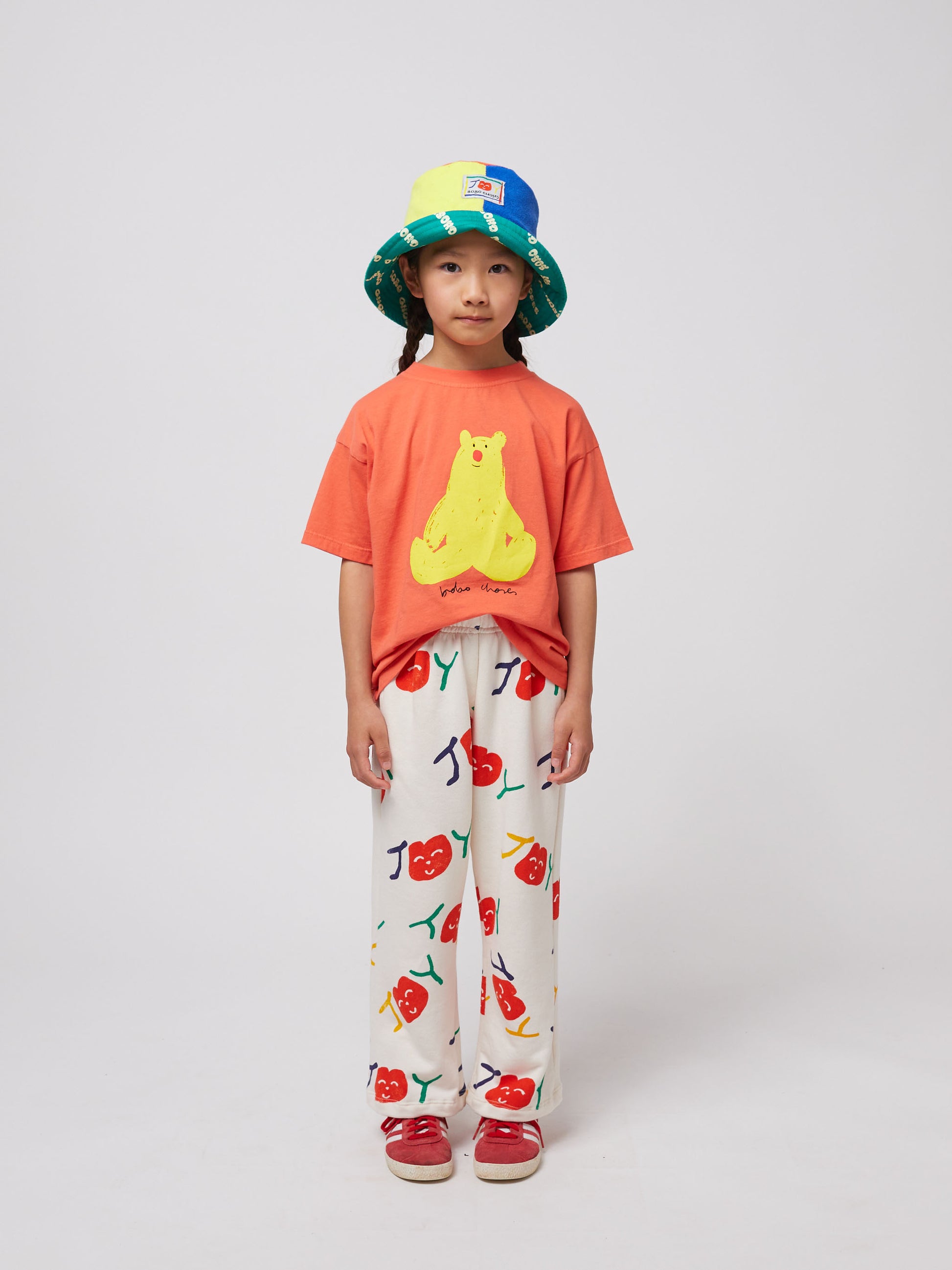 The child is standing against a plain background, dressed in the coral T-shirt, wide-leg cream trousers with a playful cherry print, and red sneakers.