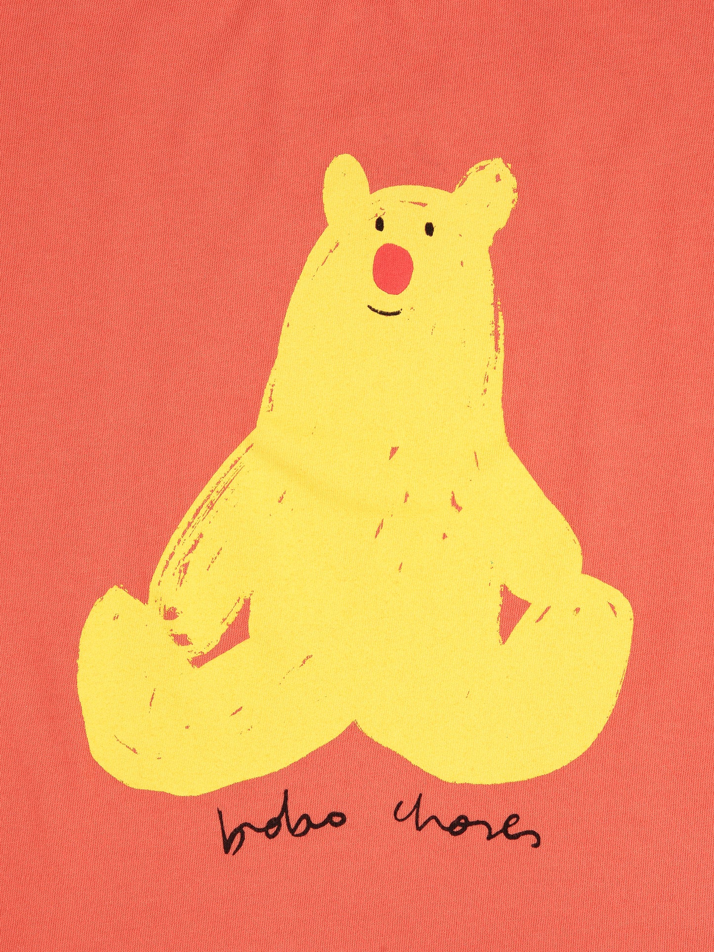 A detailed view of the yellow bear graphic on the coral T-shirt. The bear has a minimalistic, hand-drawn appearance with a round red nose, small black eyes, and a slight smile. Below the bear, "Bobo Choses" is printed in a playful, handwritten-style font.