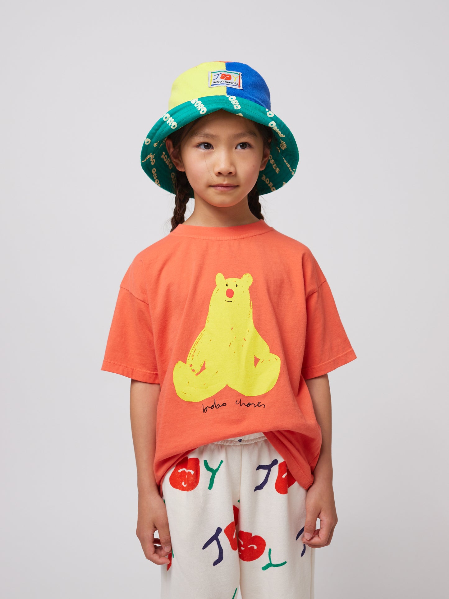 A young child with two braids is wearing the coral T-shirt with the yellow bear print. The child is also wearing a colourful bucket hat with blue, green, and yellow panels.