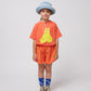A child wears the coral Bobo Choses T-shirt featuring a yellow bear print, paired with textured orange shorts with a small embroidered sunflower detail. They complete the look with a denim bucket hat, blue socks with "Bobo Choses" text, and well-worn high-top sneakers.