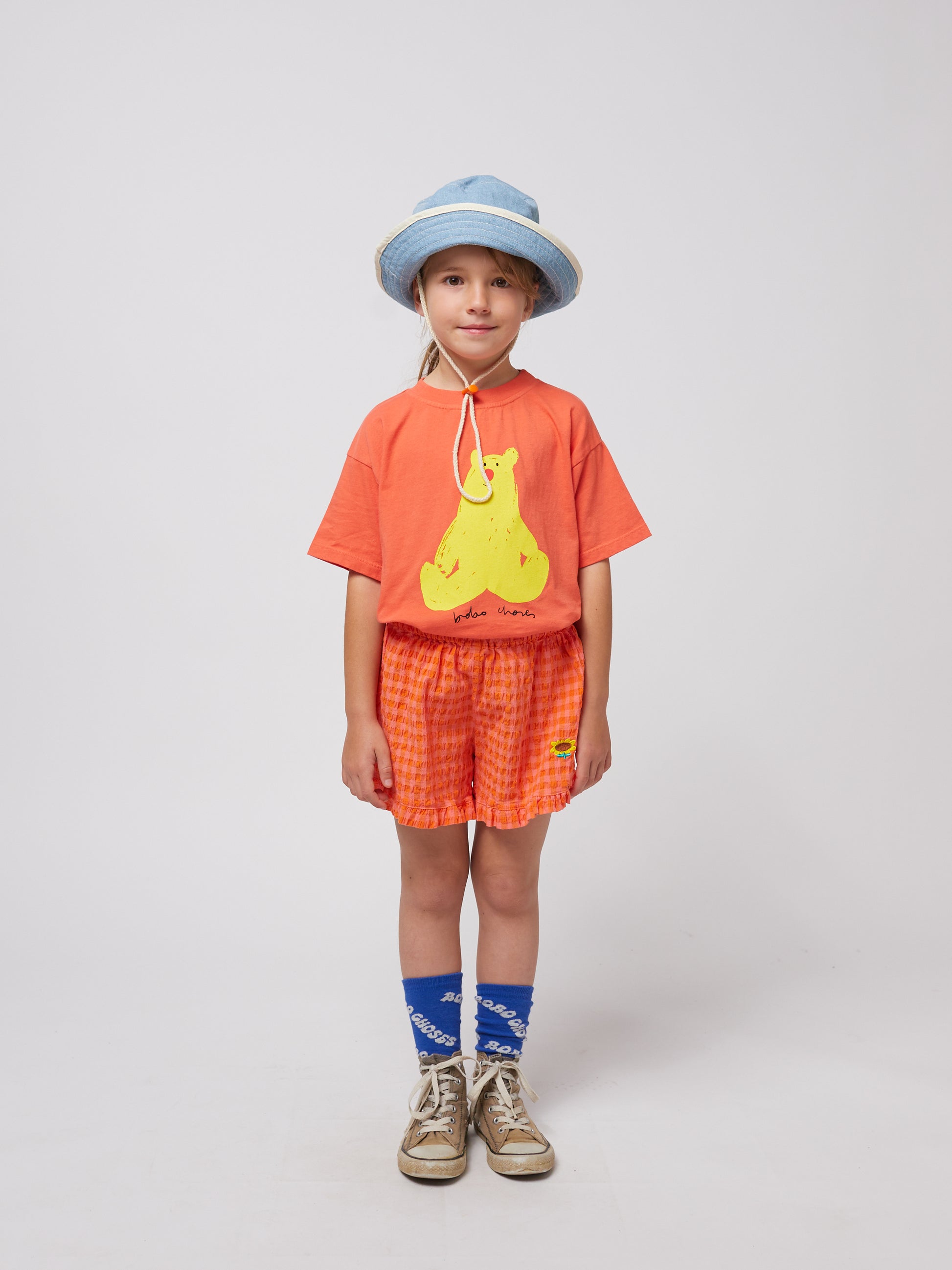 A child wears the coral Bobo Choses T-shirt featuring a yellow bear print, paired with textured orange shorts with a small embroidered sunflower detail. They complete the look with a denim bucket hat, blue socks with "Bobo Choses" text, and well-worn high-top sneakers.