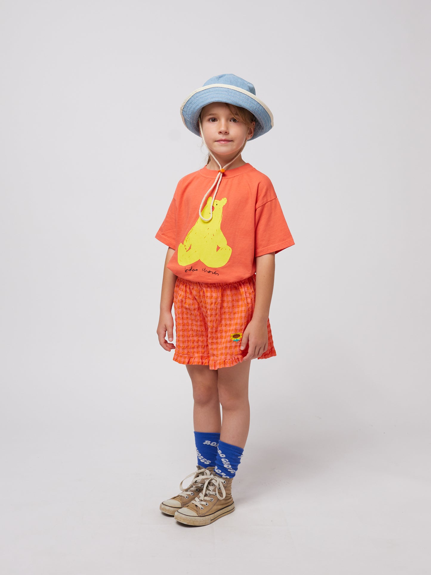 A child wearing the coral T-shirt is captured from the side, paired with textured orange shorts, a denim bucket hat, blue socks, and vintage-style sneakers.