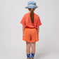 A child stands facing away from the camera, wearing the coral T-shirt with orange textured shorts. They wear a denim bucket hat and blue socks with "Bobo Choses" branding.