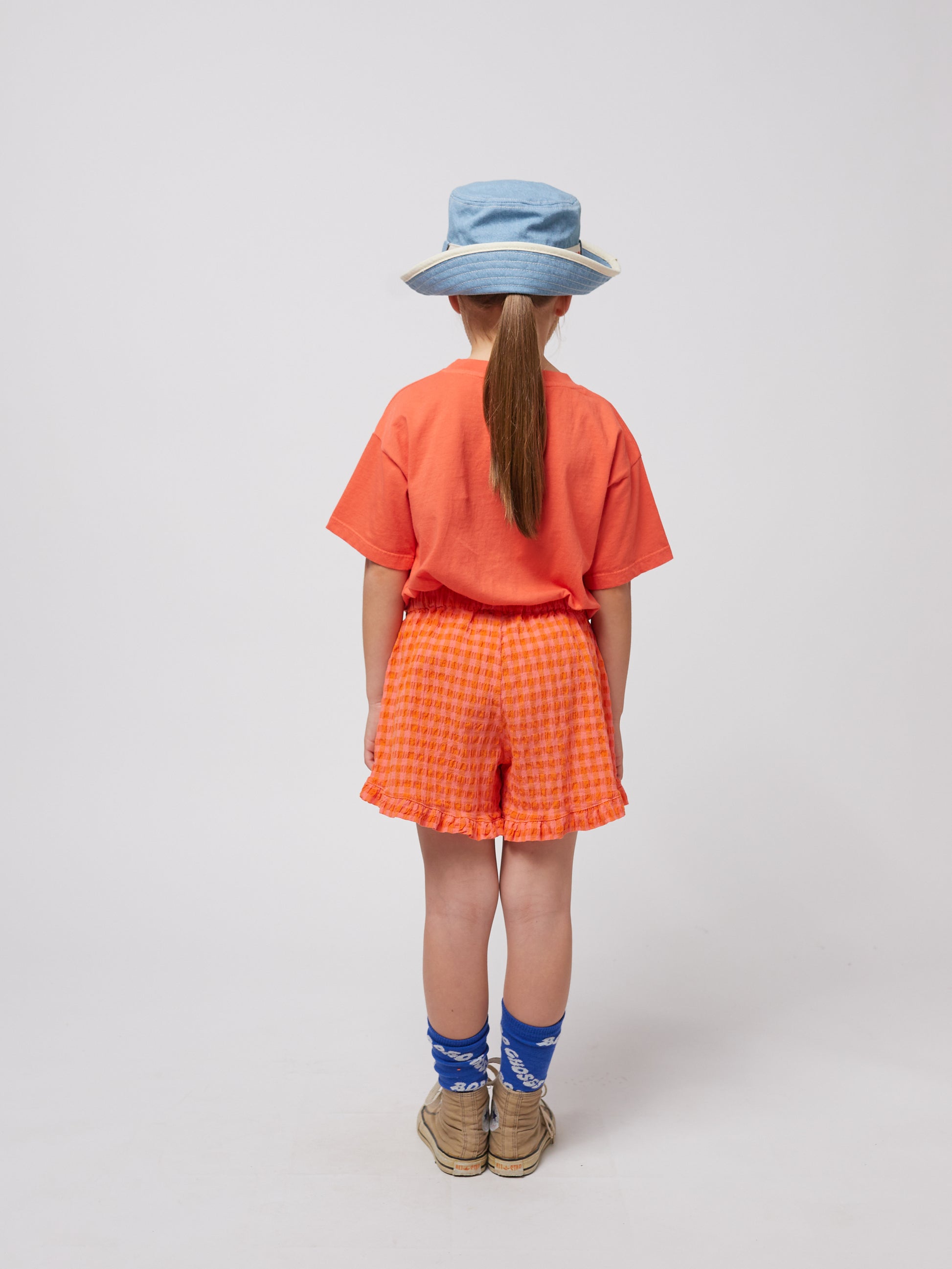 A child stands facing away from the camera, wearing the coral T-shirt with orange textured shorts. They wear a denim bucket hat and blue socks with "Bobo Choses" branding.