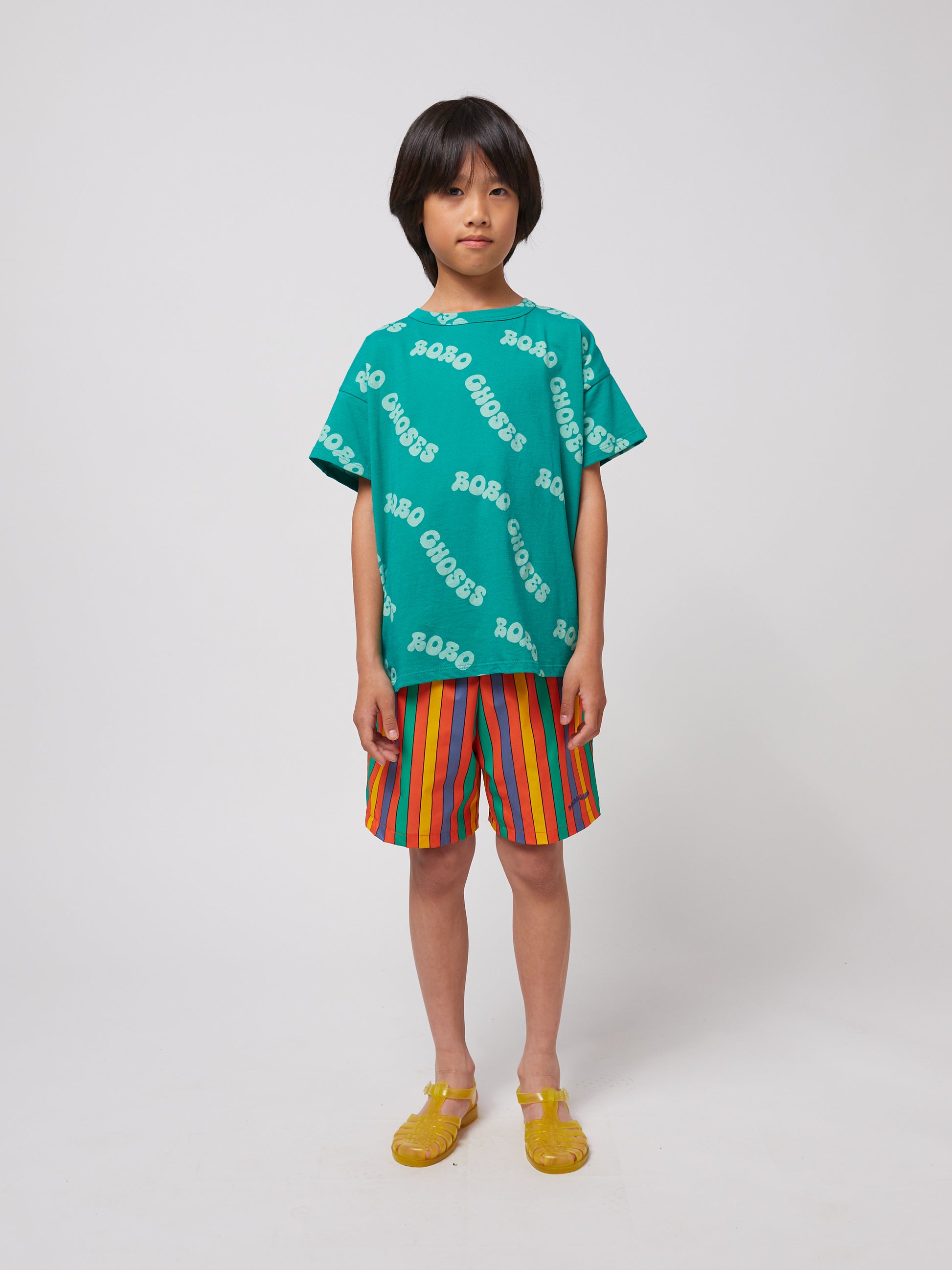 A relaxed-fit green "BOBO CHOSES" T-shirt is paired with bright, vertical-striped shorts and yellow jelly sandals.