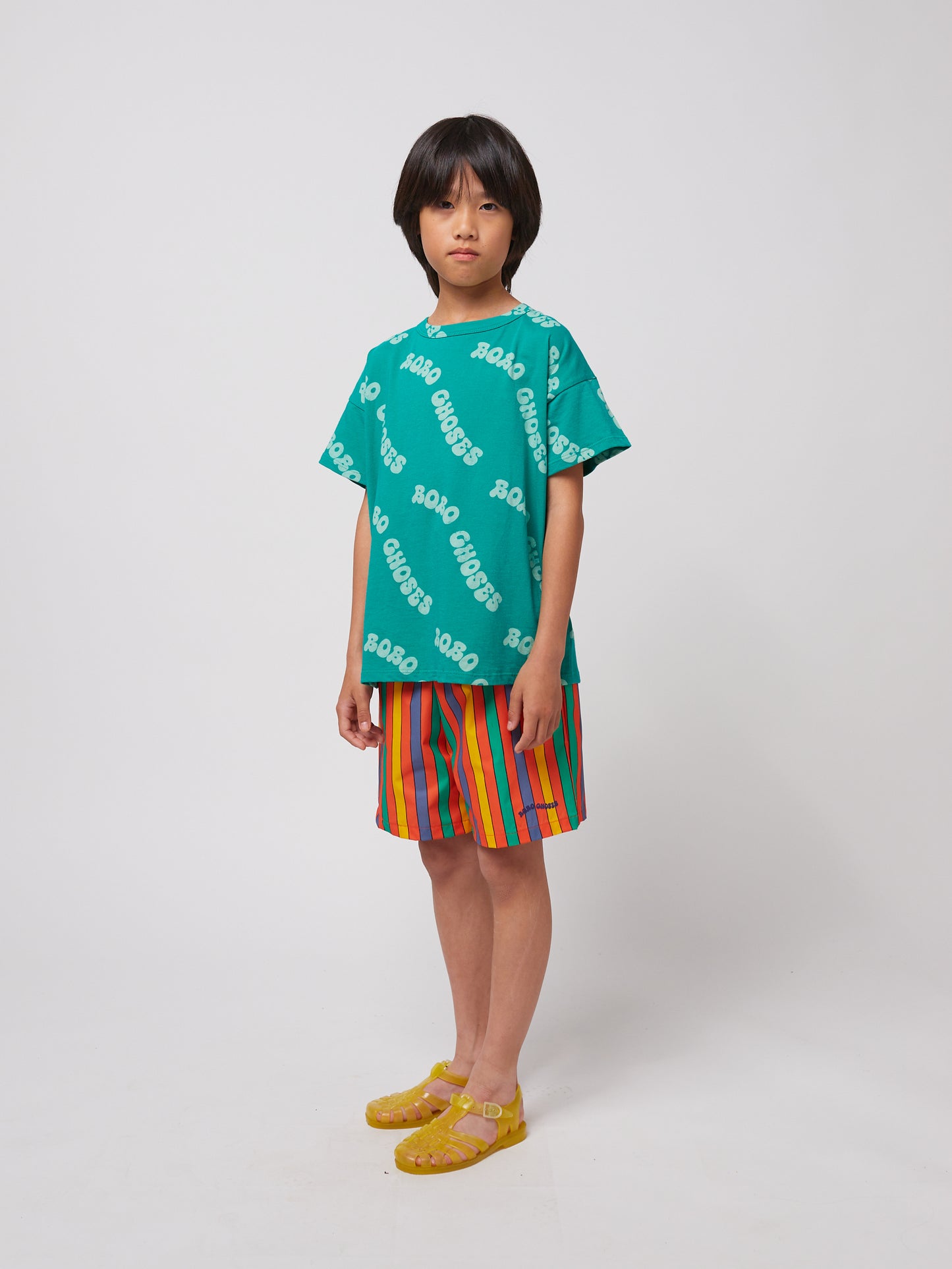 A child stands wearing the green "BOBO CHOSES" all-over print T-shirt, paired with bold multicoloured striped shorts and translucent yellow jelly sandals.