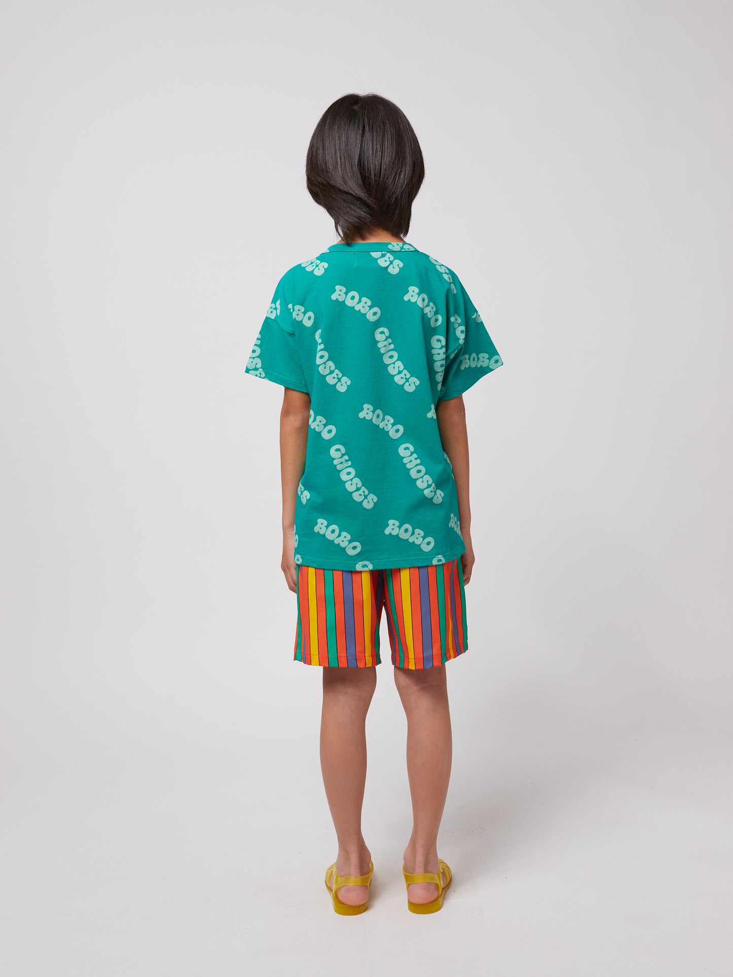 A child is seen from behind, wearing the green T-shirt with bright multicoloured striped shorts and yellow sandals.