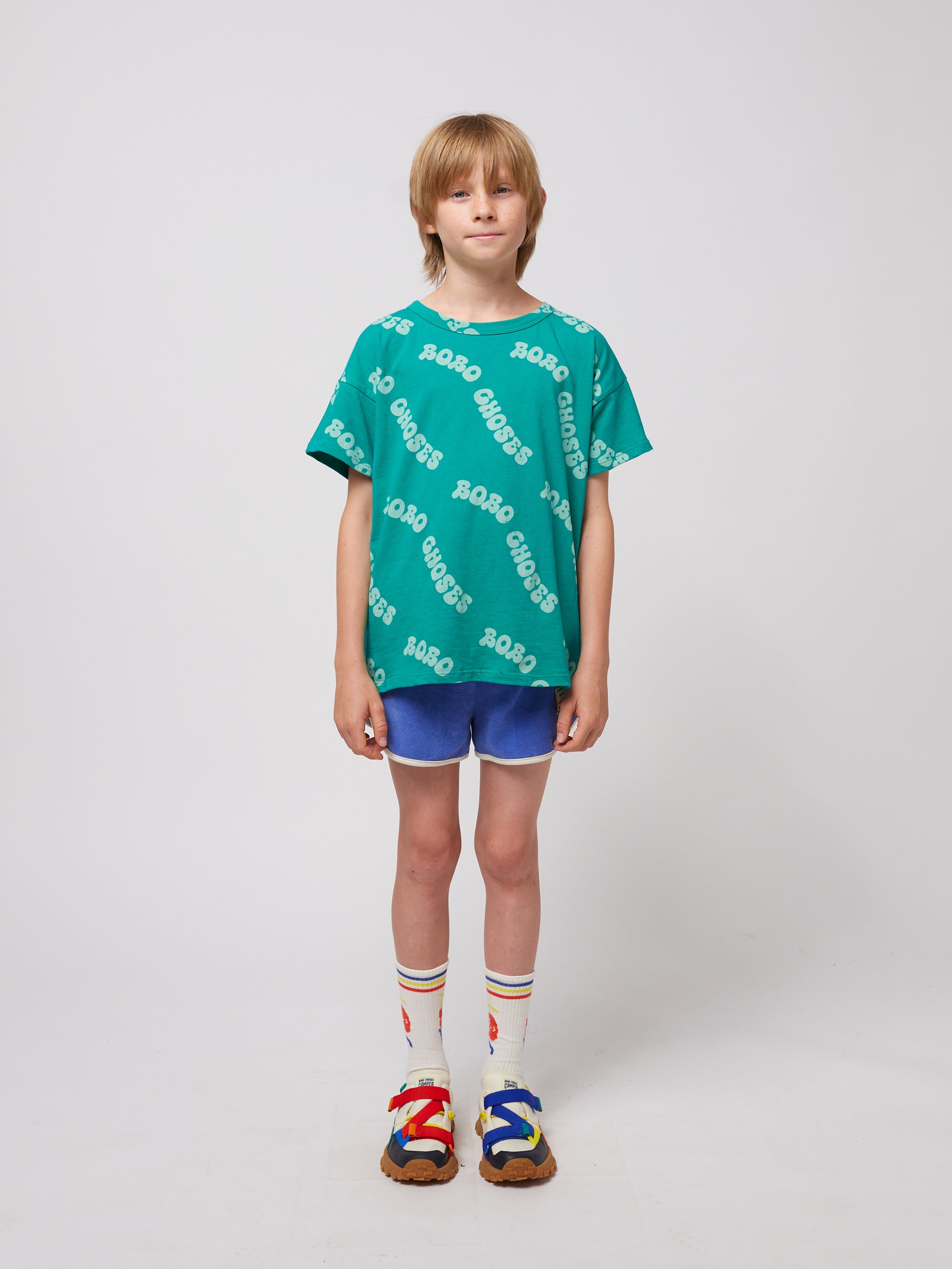 A child stands wearing the green "BOBO CHOSES" T-shirt, paired with blue athletic-style shorts, cream socks with multicoloured stripes, and vibrant strappy sandals.