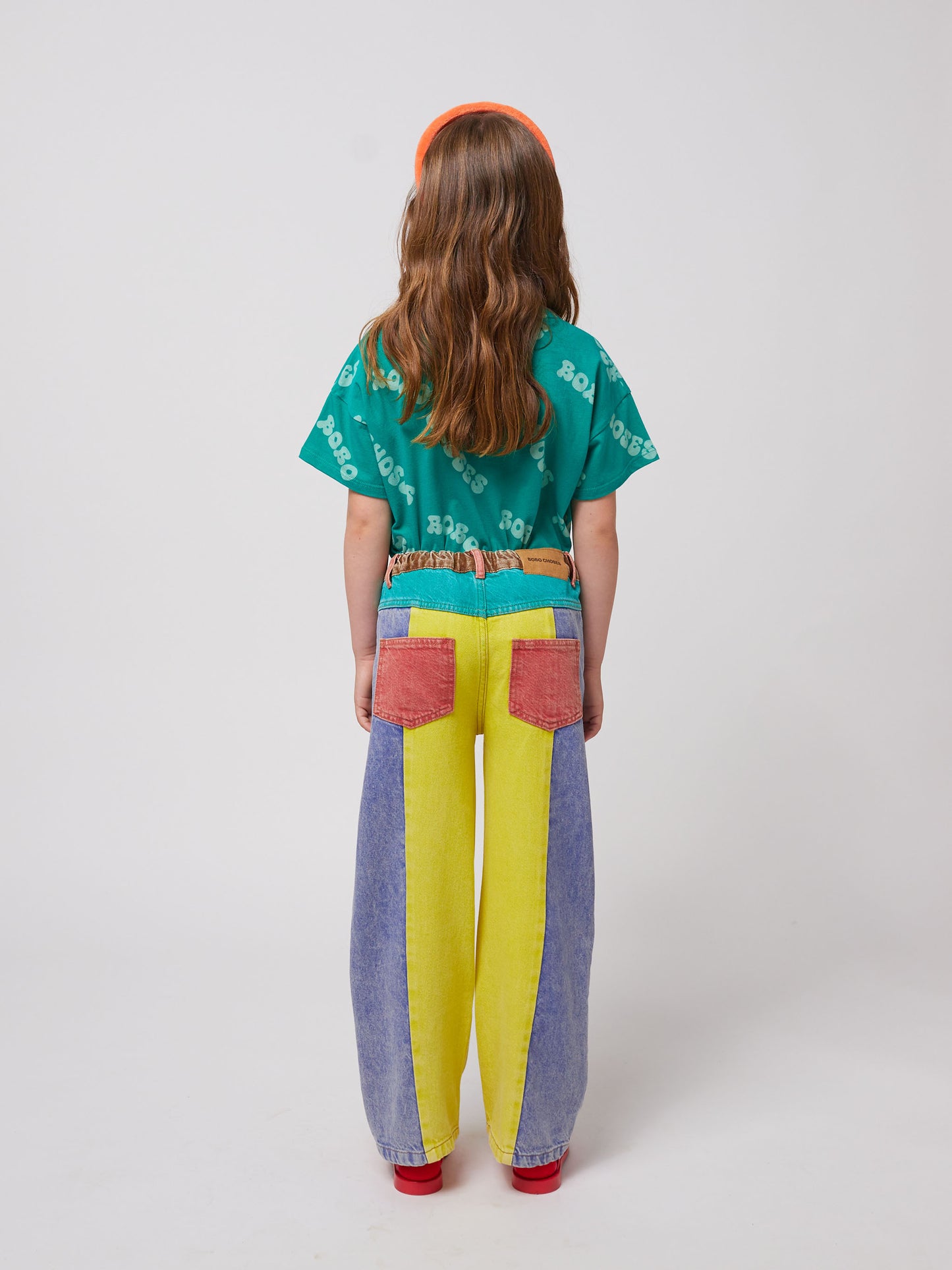 A child with long, wavy brown hair is seen from behind wearing the green T-shirt, paired with high-waisted wide-leg jeans featuring bold yellow, blue, and red colour-blocking.