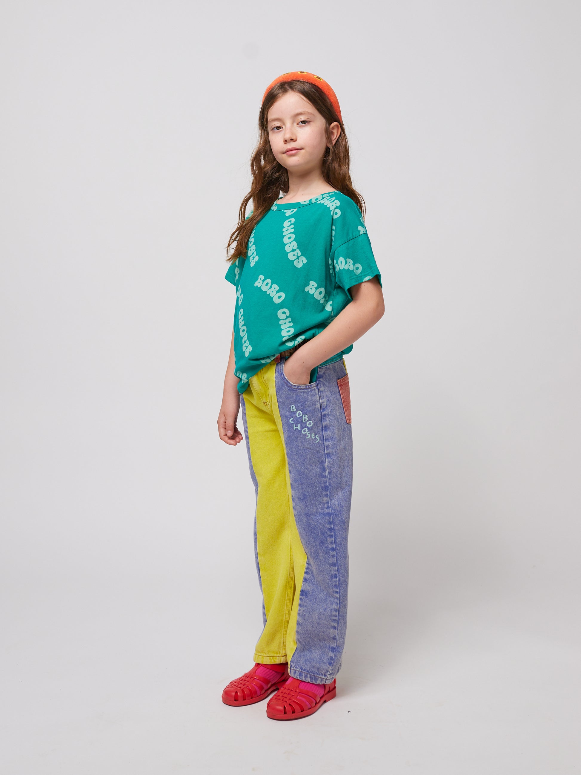 A child wears the oversized green "BOBO CHOSES" T-shirt, tucked into colour-blocked denim jeans in shades of yellow, blue, and red. The outfit is finished with an orange headband and red jelly sandals.