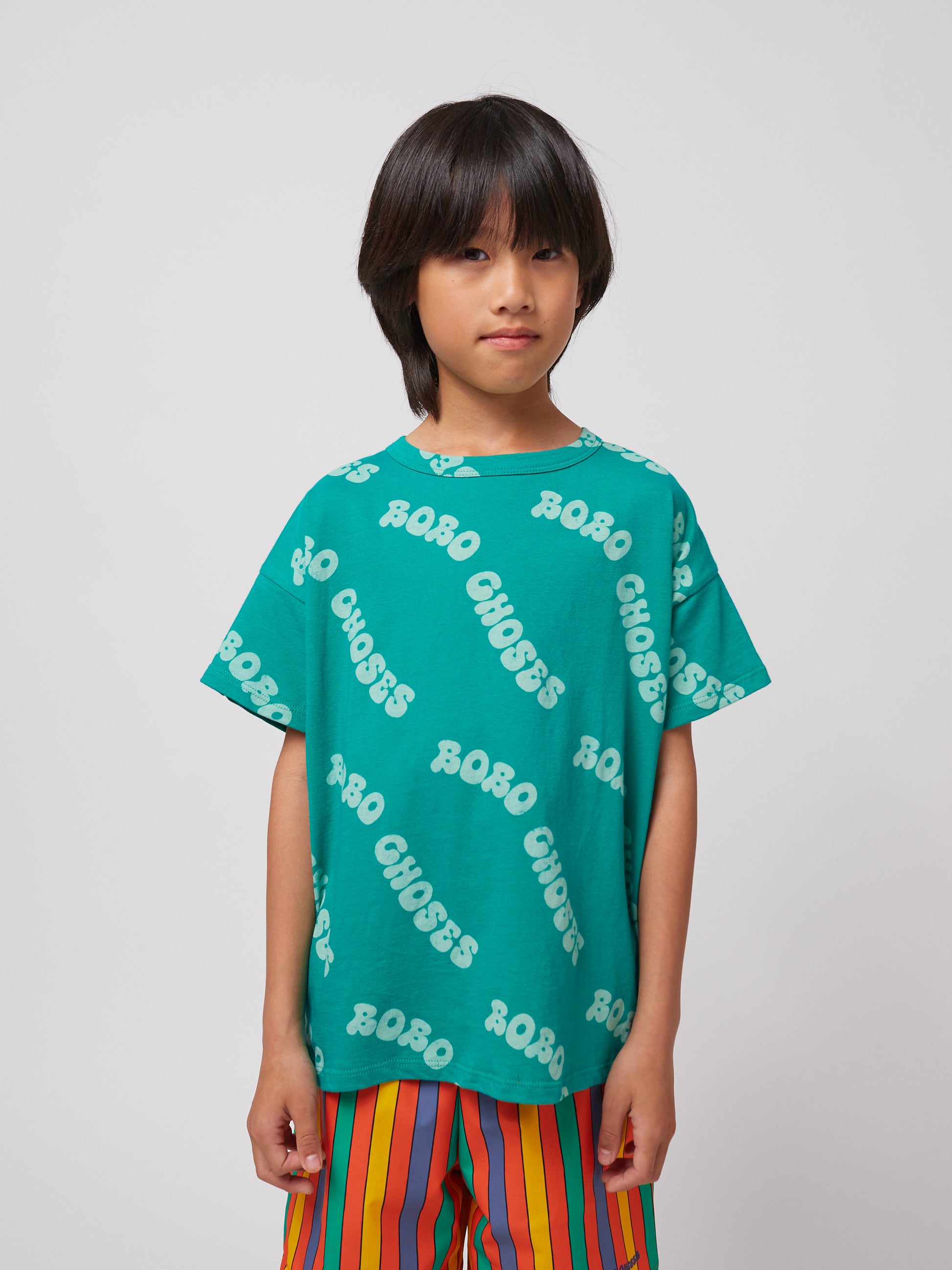 A child models the green T-shirt with "BOBO CHOSES" text scattered across it, along with brightly striped shorts in red, blue, green, and yellow.