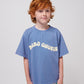 A young child with curly red hair and freckles wears a blue Bobo Choses Wavy T-Shirt with a cream-coloured wavy logo across the chest. They pair the oversized T-shirt with patterned shorts in white and green. The child has a soft smile and a relaxed posture, standing against a plain white background.