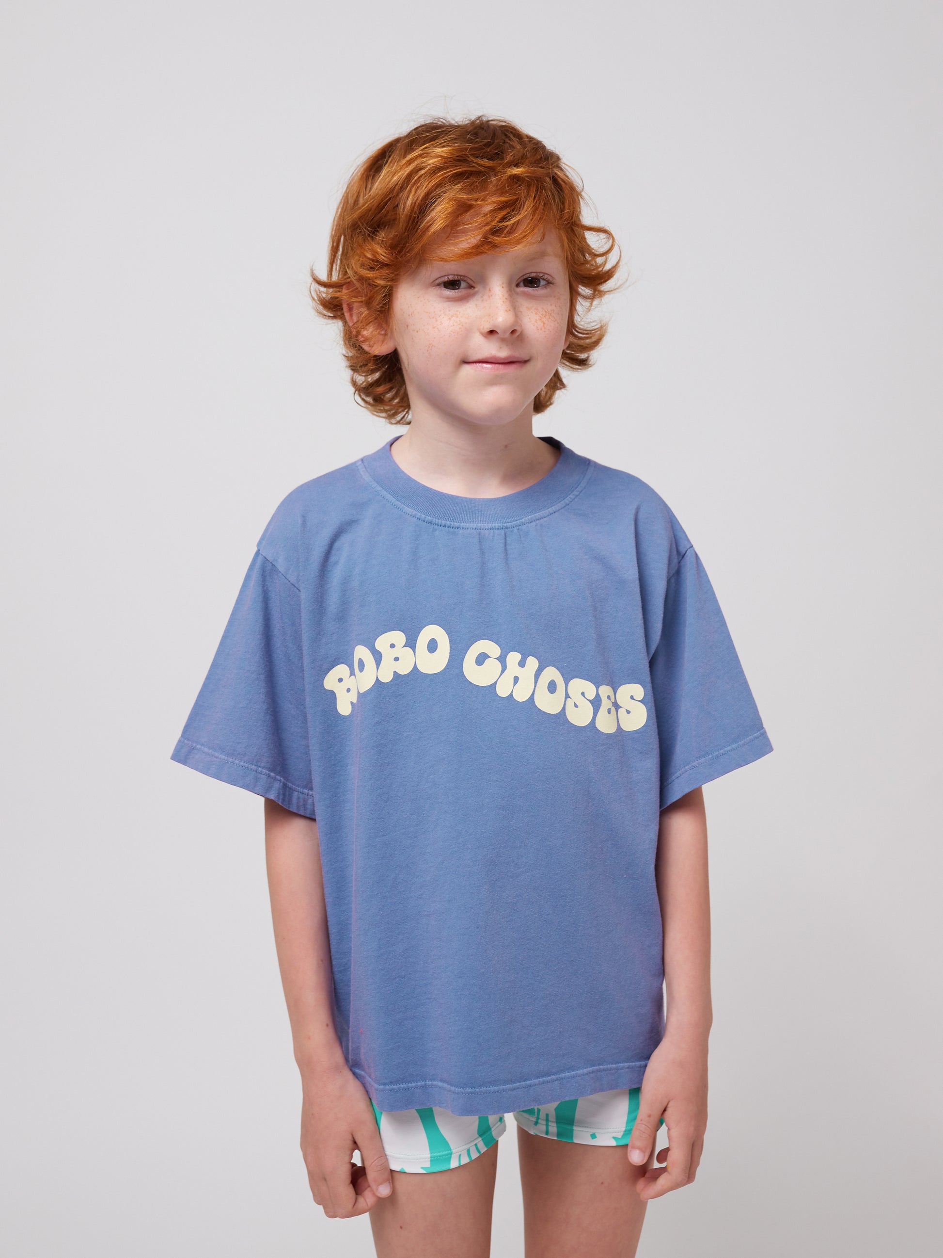 A young child with curly red hair and freckles wears a blue Bobo Choses Wavy T-Shirt with a cream-coloured wavy logo across the chest. They pair the oversized T-shirt with patterned shorts in white and green. The child has a soft smile and a relaxed posture, standing against a plain white background.