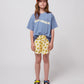 Girl wearing the blue wavy t-shirt tucked into yellow sunflower shorts, paired with colourful sandals and a cap.