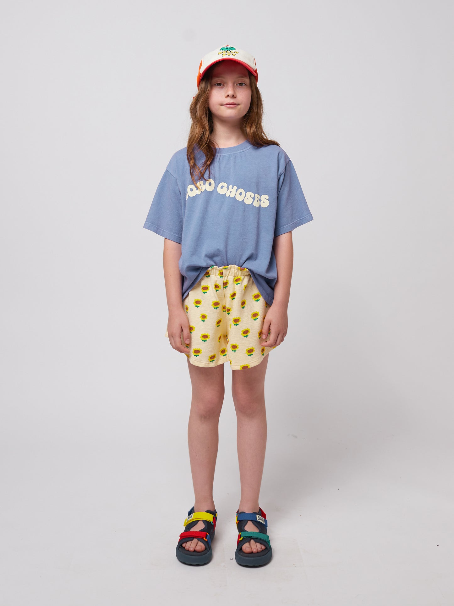 Girl wearing the blue wavy t-shirt tucked into yellow sunflower shorts, paired with colourful sandals and a cap.