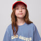 A young girl with long, wavy brown hair wears a blue Bobo Choses Wavy T-Shirt featuring a cream-coloured wavy logo across the chest. She also wears a red and white cap with an embroidered design, looking directly at the camera with a relaxed expression. The close-up shot highlights the texture of the T-shirt and the playful typography of the logo.