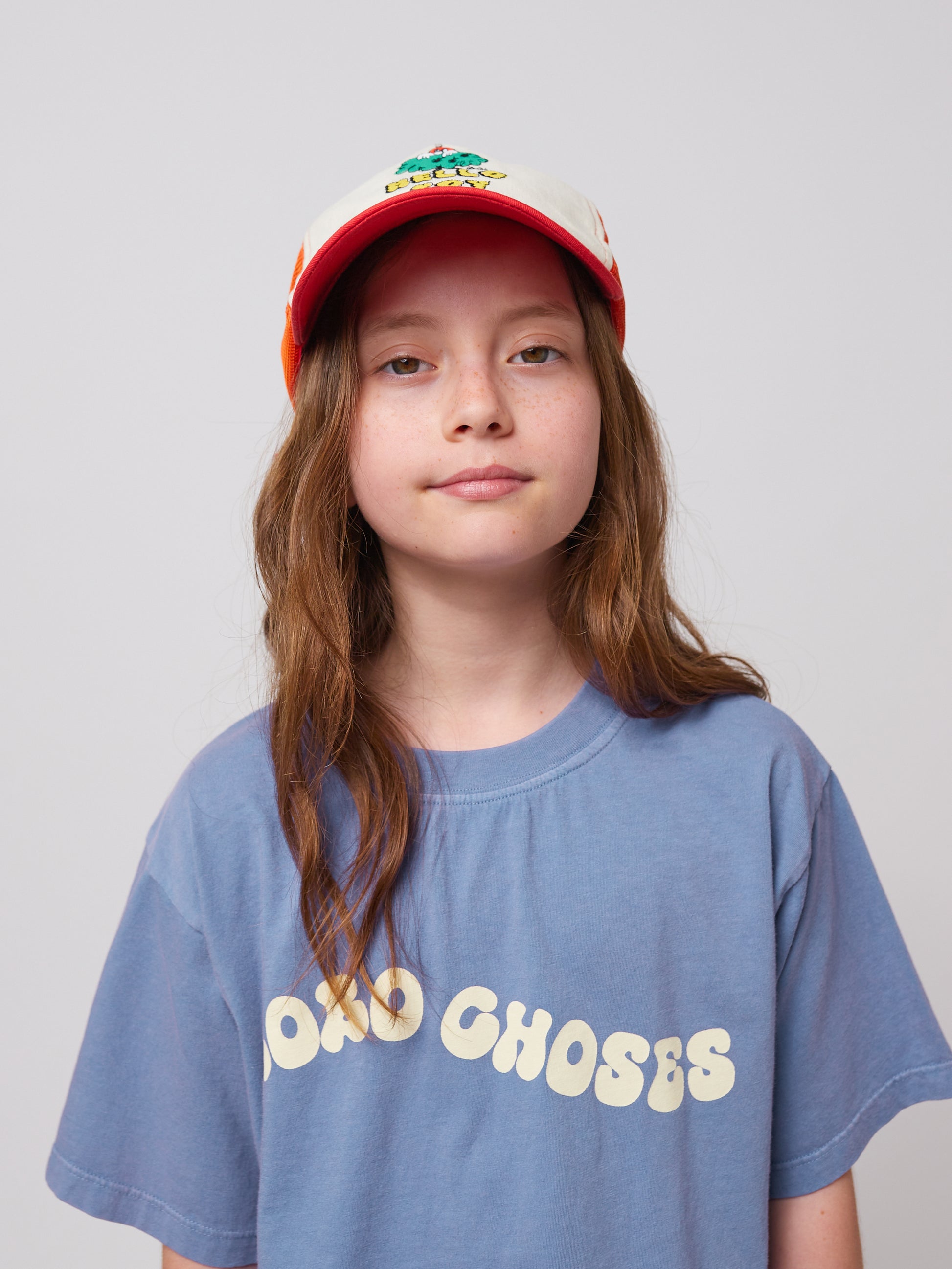 A young girl with long, wavy brown hair wears a blue Bobo Choses Wavy T-Shirt featuring a cream-coloured wavy logo across the chest. She also wears a red and white cap with an embroidered design, looking directly at the camera with a relaxed expression. The close-up shot highlights the texture of the T-shirt and the playful typography of the logo.