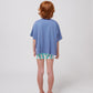 A young child with curly red hair stands facing away from the camera, showcasing the back of a loose-fitting blue Bobo Choses Wavy T-Shirt. The shirt drapes over patterned green and white shorts, and they wear red jelly sandals. The background is plain white.