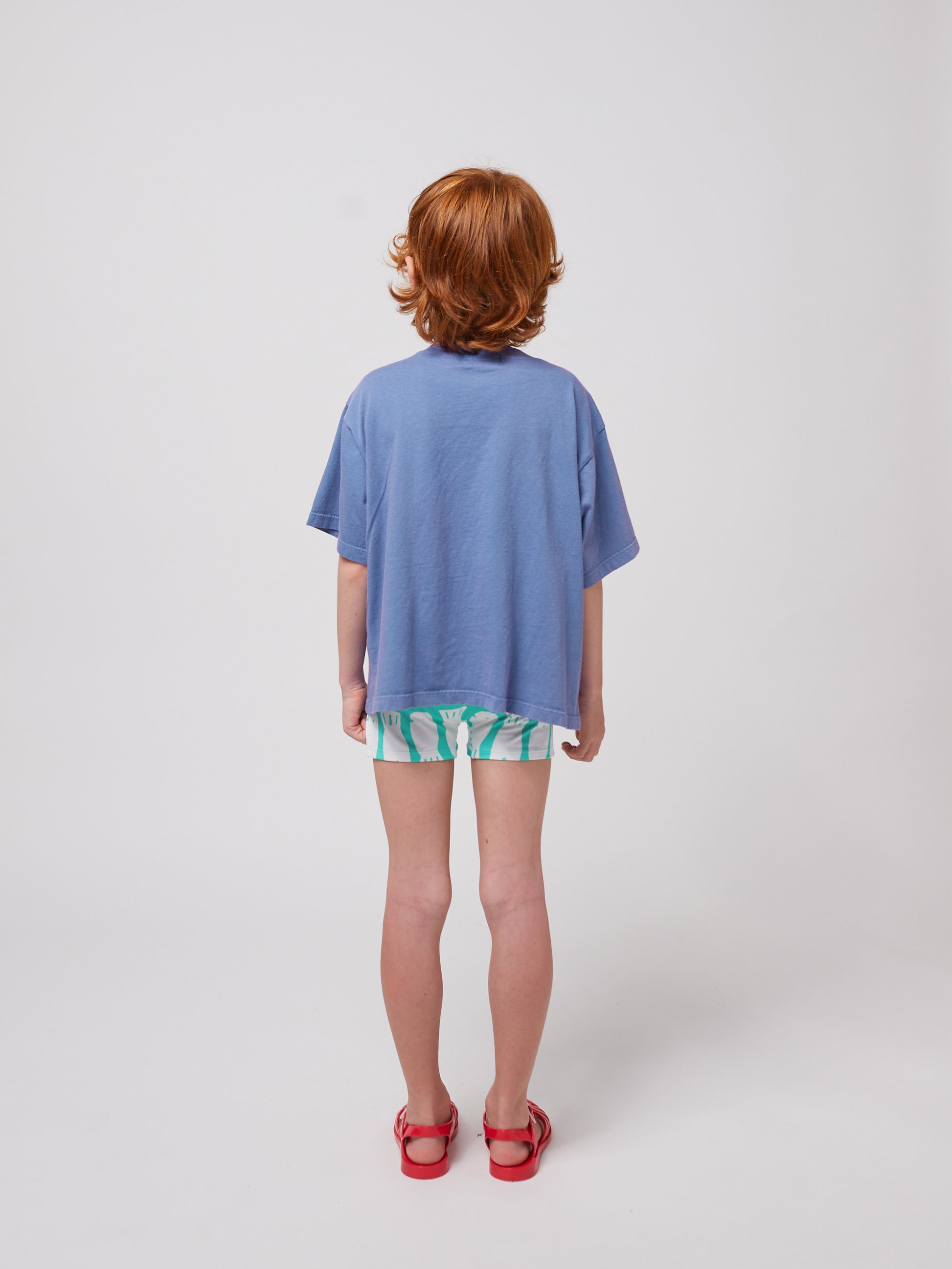 A young child with curly red hair stands facing away from the camera, showcasing the back of a loose-fitting blue Bobo Choses Wavy T-Shirt. The shirt drapes over patterned green and white shorts, and they wear red jelly sandals. The background is plain white.