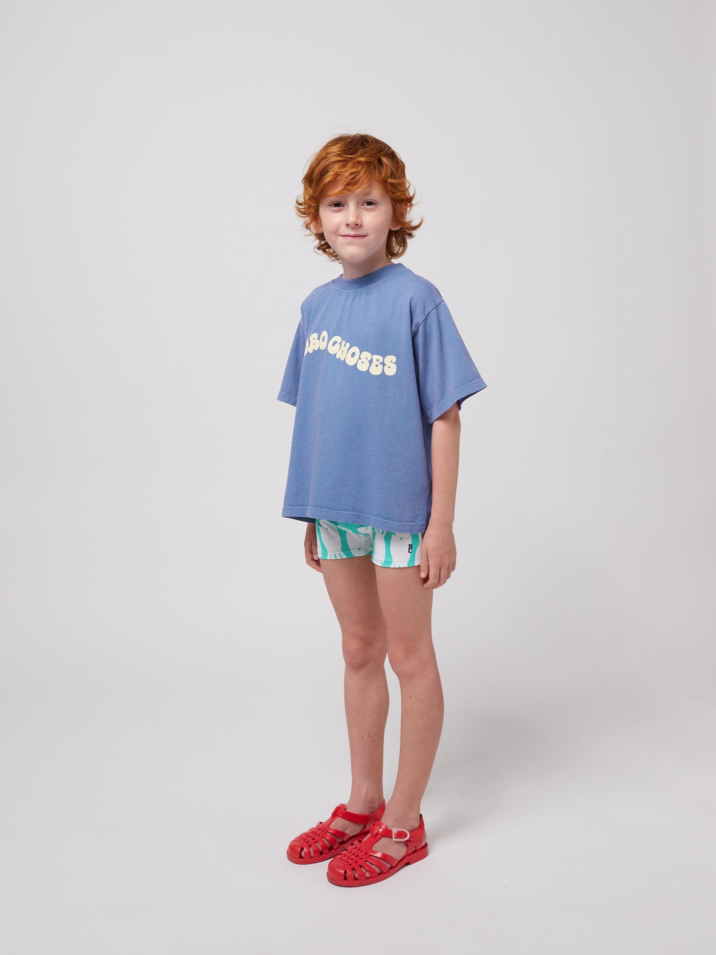 A young child with curly red hair and freckles wears a loose-fitting blue Bobo Choses Wavy T-Shirt with a cream wavy logo across the chest. They pair it with patterned green and white shorts and red jelly sandals. The child stands slightly angled with a soft smile against a plain white background.