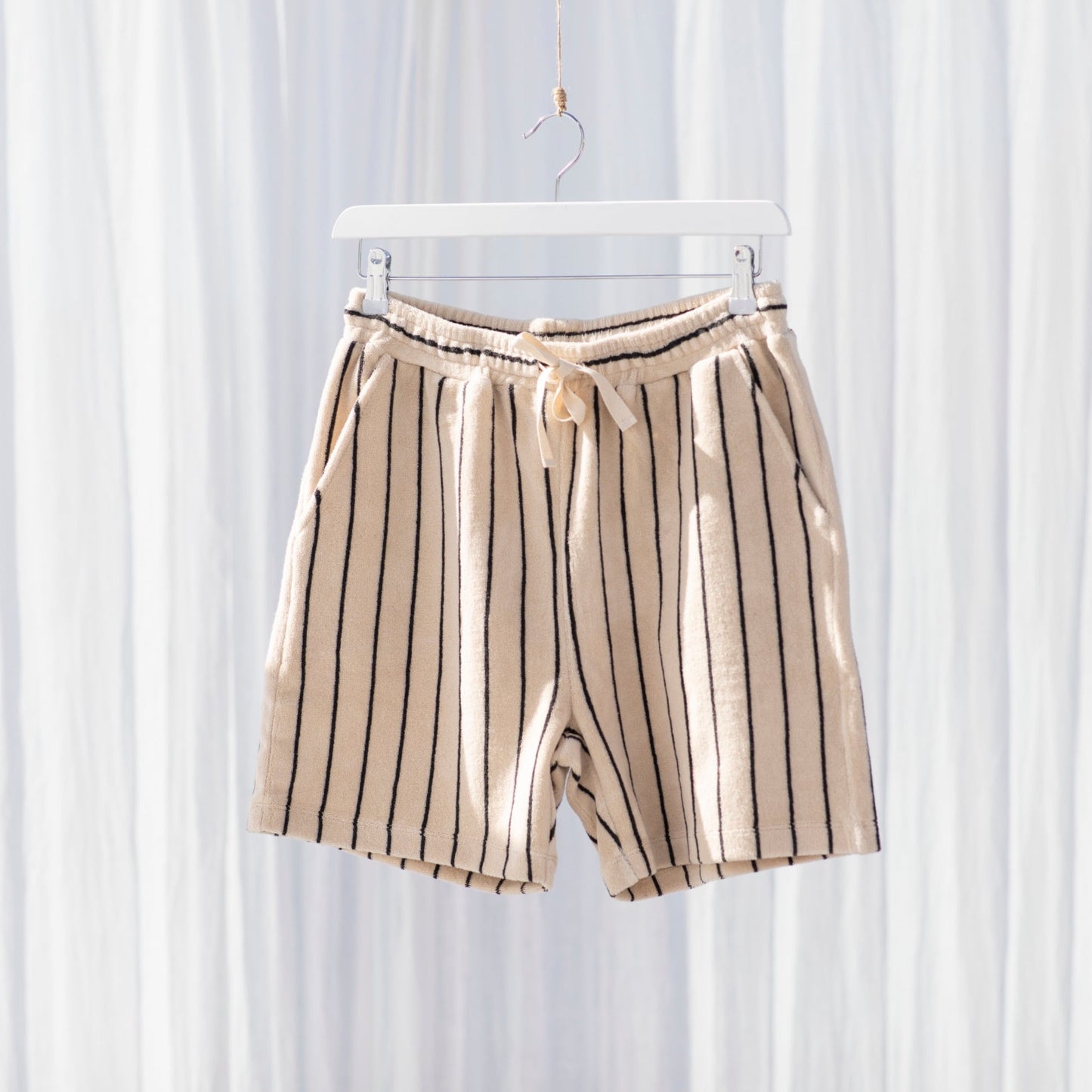 A pair of cream and thinly black striped above the knee shorts with a drawstring and elasticated waistband hanging on a white hanger in front of a white curtain. 