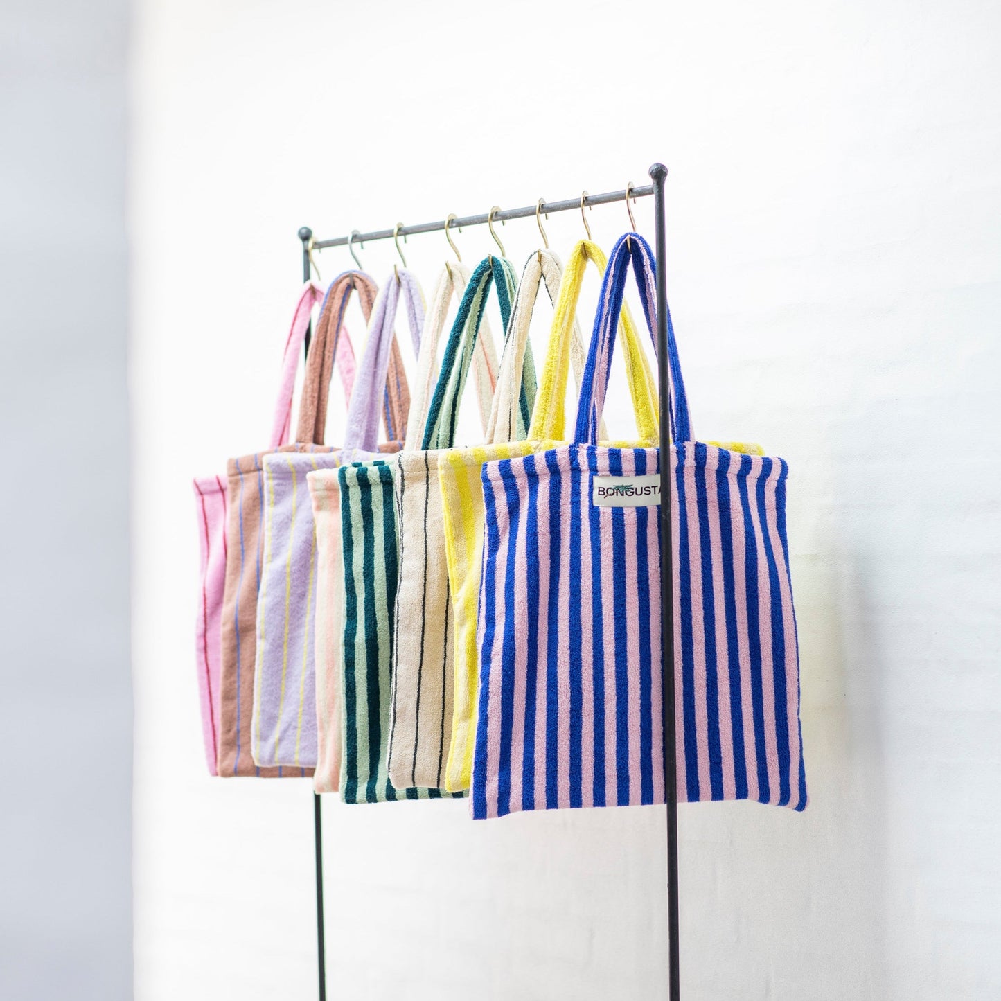 8 Bongusta classic tote bags hang on rustic gold hangers on a black rail. The bags are pink and thin neon striped, brown and thin blue striped, lilac and thin neon yellow striped, cream and pink striped, light and dark green striped, cream and thin black striped, cream and neon yellow striped and a pink and neon blue striped bag. 