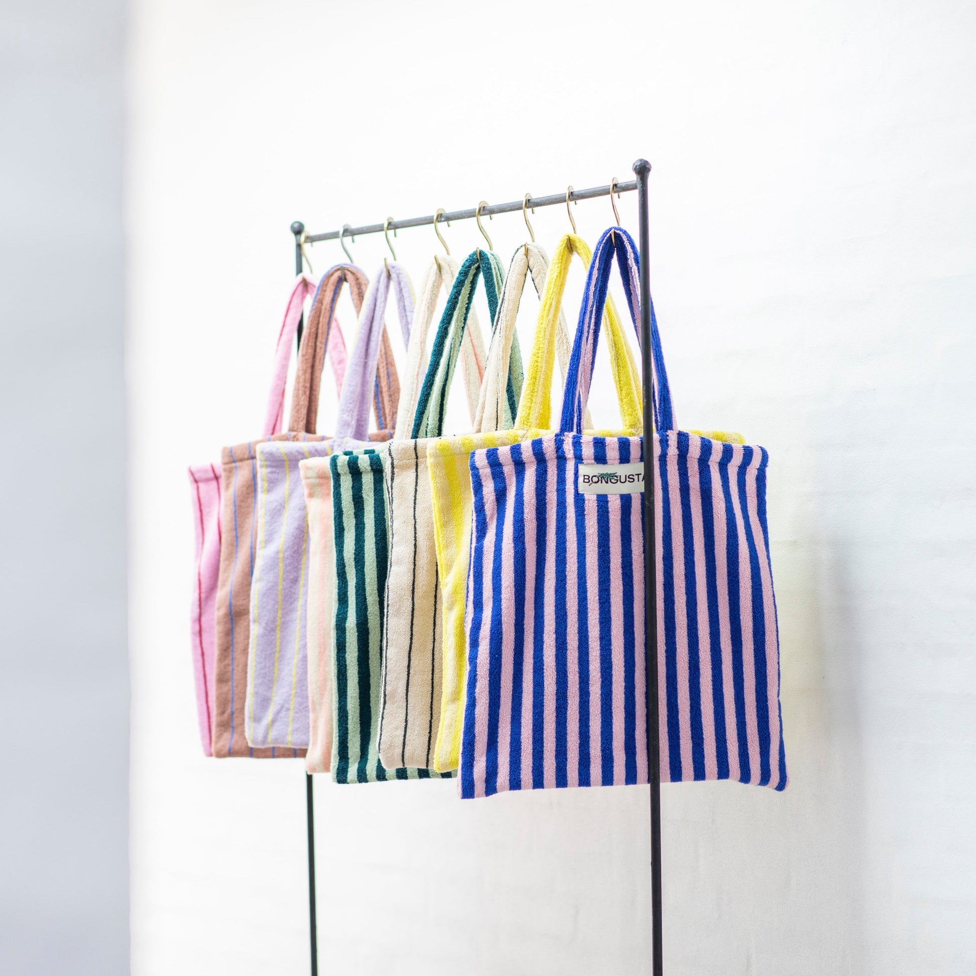 8 Bongusta classic tote bags hang on rustic gold hangers on a black rail. The bags are pink and thin neon striped, brown and thin blue striped, lilac and thin neon yellow striped, cream and pink striped, light and dark green striped, cream and thin black striped, cream and neon yellow striped and a pink and neon blue striped bag. 