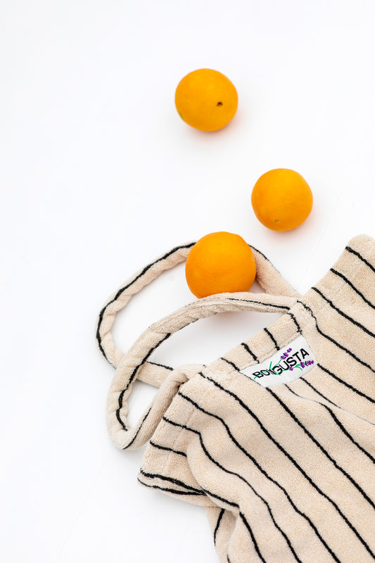 A cream and thin black striped classic tote bag, made of towelling fabric with a  cream and thin black striped handle laying on a white floor with 3 oranges rolling out of the bag. At the top of the bag it says BONGUSTA in black inside of a cream box with green and lilac flowers coming off the words bongusta. 