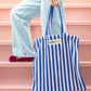 A baby pink and deep blue striped classic tote bag, made of towelling fabric with a baby pink and deep blue striped handle is held by someone standing on pink steps wearing pin, velvet flats with a bow on them, blue wide leg jeans and a cream and thinly black striped button up shirt, also made of towelling fabric. At the top of the bag it says BONGUSTA in black inside of a cream box with green and lilac flowers coming off the words bongusta. 
