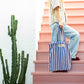 A baby pink and deep blue striped classic tote bag, made of towelling fabric with a baby pink and deep blue striped handle is held by someone standing on pink steps, with a large green cactus next to the steps, wearing pin, velvet flats with a bow on them, blue wide leg jeans and a cream and thinly black striped button up shirt, also made of towelling fabric. At the top of the bag it says BONGUSTA in black inside of a cream box with green and lilac flowers coming off the words bongusta. 