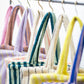 8 Bongusta classic tote bags hang on rustic gold hangers on a black rail, with only the handles visible. The bags are pink and thin neon striped, brown and thin blue striped, lilac and thin neon yellow striped, cream and pink striped, light and dark green striped, cream and thin black striped, cream and neon yellow striped and a pink and neon blue striped bag. 