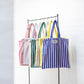 8 Bongusta classic tote bags hang on rustic gold hangers on a black rail. The bags are pink and thin neon striped, brown and thin blue striped, lilac and thin neon yellow striped, cream and pink striped, light and dark green striped, cream and thin black striped, cream and neon yellow striped and a pink and neon blue striped bag. 