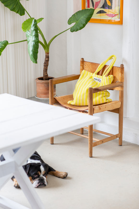 A cream and neon yellow striped classic tote bag, made of towelling fabric with a cream and neon yellow striped handle sits on top of a wooden chair next to a green potted plant, in front of a white table with a dog laying on the floor and above the chair a print. At the top of the bag it says BONGUSTA in black inside of a cream box with green and lilac flowers coming off the words bongusta. 