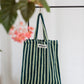 A light and dark green striped classic tote bag, made of towelling fabric with a long light and dark green striped handle is hanging off a rustic gold hanger on a black rail with a green plant at the front of the image. At the top of the bag it says BONGUSTA in black inside of a cream box with green flowers coming off the words bongusta. 