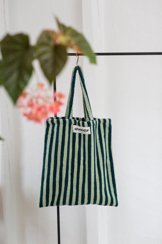 A light and dark green striped classic tote bag, made of towelling fabric with a long light and dark green striped handle is hanging off a rustic gold hanger on a black rail with a green plant at the front of the image. At the top of the bag it says BONGUSTA in black inside of a cream box with green flowers coming off the words bongusta. 