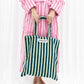 A light and dark green striped classic tote bag, made of towelling fabric with a long light and dark green striped handle is held by someone with both hands in front of them, wearing no shoes and a pink and thinly striped neon red towelling dressing gown.  At the top of the bag it says BONGUSTA in black inside of a cream box with green flowers coming off the words bongusta. 