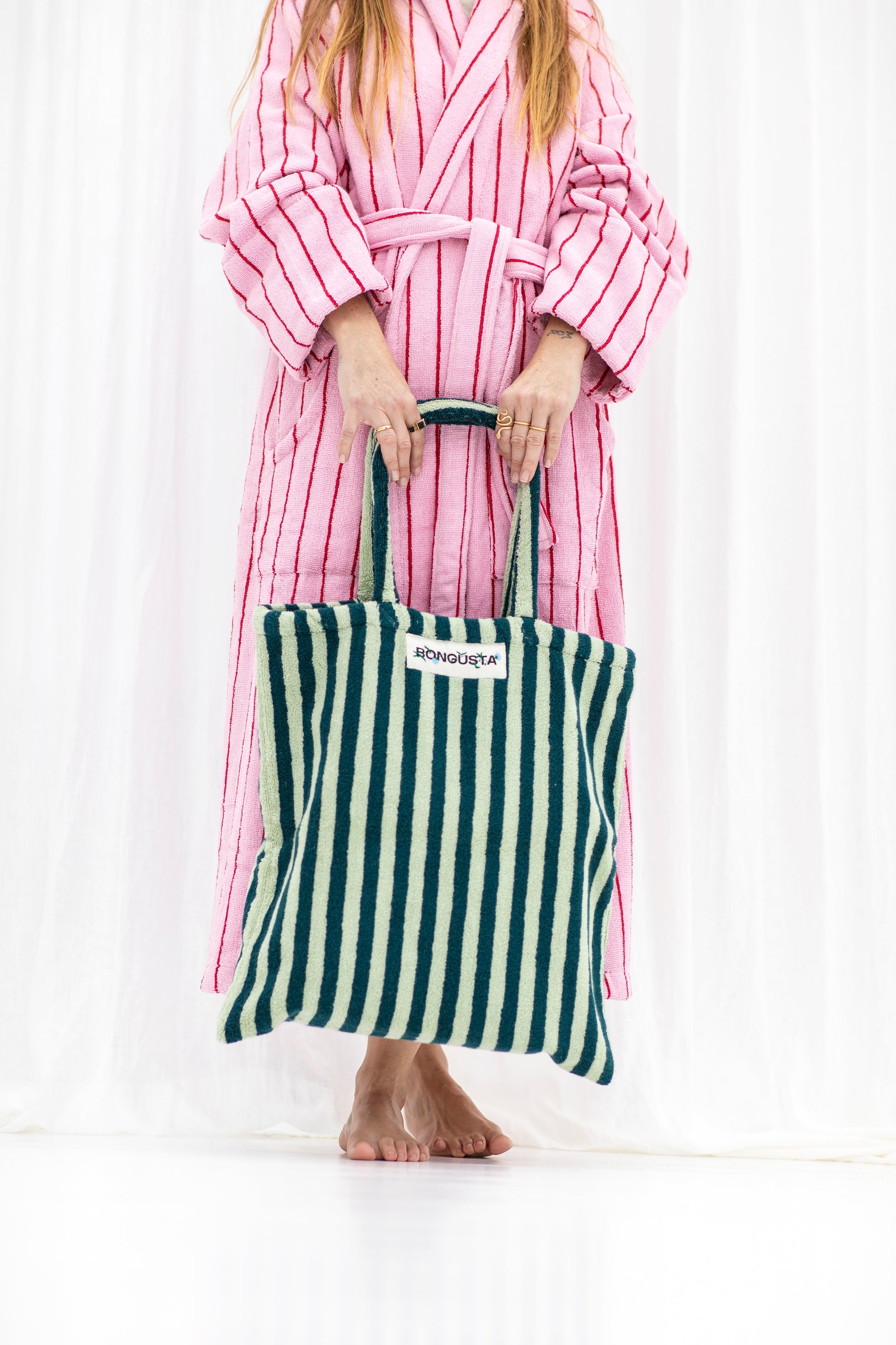 A light and dark green striped classic tote bag, made of towelling fabric with a long light and dark green striped handle is held by someone with both hands in front of them, wearing no shoes and a pink and thinly striped neon red towelling dressing gown.  At the top of the bag it says BONGUSTA in black inside of a cream box with green flowers coming off the words bongusta. 