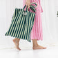 A light and dark green striped classic tote bag, made of towelling fabric with a long light and dark green striped handle is held in one hand by someone walking past a white curtain, wearing no shoes and a pink and thinly striped neon red towelling dressing gown.  At the top of the bag it says BONGUSTA in black inside of a cream box with green flowers coming off the words bongusta. 