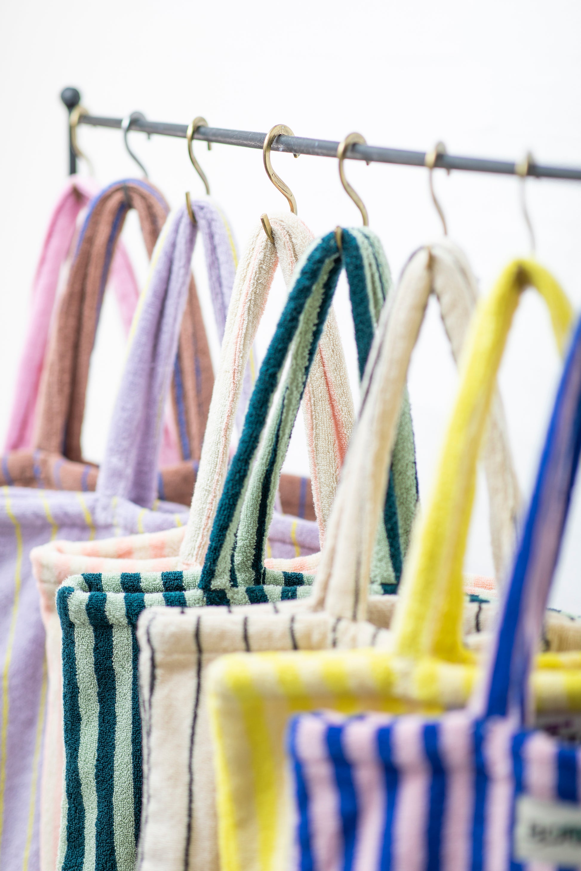 8 Bongusta classic tote bags hang on rustic gold hangers on a black rail, with only the handles visible. The bags are pink and thin neon striped, brown and thin blue striped, lilac and thin neon yellow striped, cream and pink striped, light and dark green striped, cream and thin black striped, cream and neon yellow striped and a pink and neon blue striped bag. 