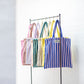 8 Bongusta classic tote bags hang on rustic gold hangers on a black rail. The bags are pink and thin neon striped, brown and thin blue striped, lilac and thin neon yellow striped, cream and pink striped, light and dark green striped, cream and thin black striped, cream and neon yellow striped and a pink and neon blue striped bag. 