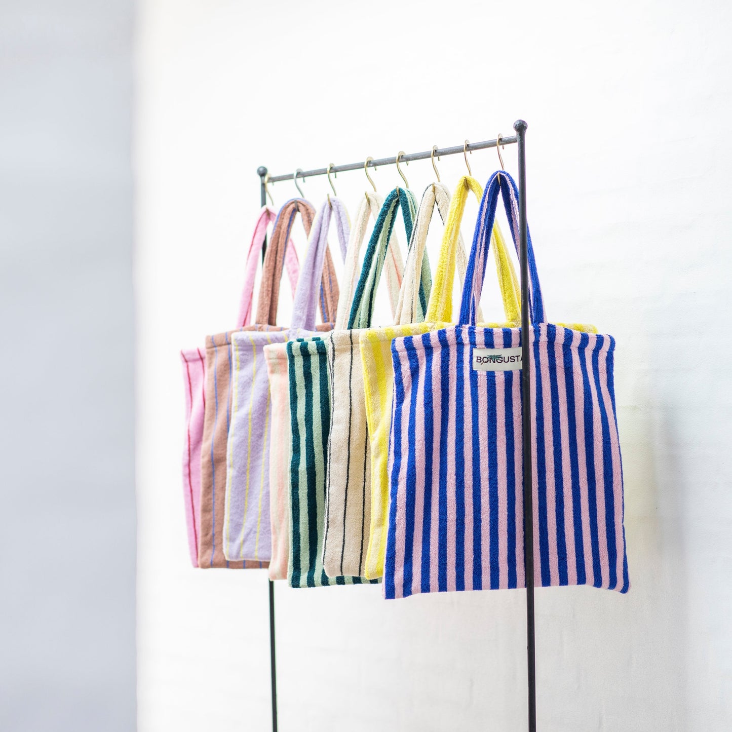 8 Bongusta classic tote bags hang on rustic gold hangers on a black rail. The bags are pink and thin neon striped, brown and thin blue striped, lilac and thin neon yellow striped, cream and pink striped, light and dark green striped, cream and thin black striped, cream and neon yellow striped and a pink and neon blue striped bag. 